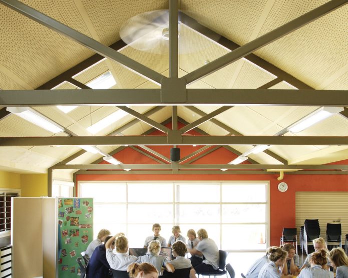 The interior of a classroom with school children sitting at tables, framed at the roof to highlight the panels installed in the roof.
